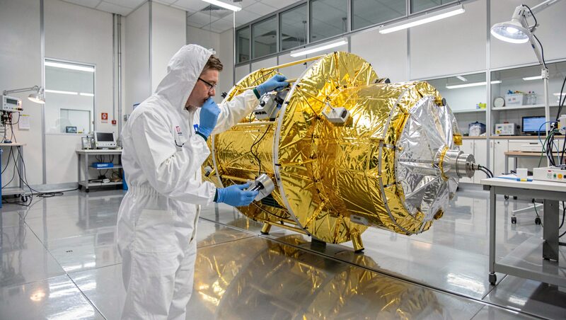 A clean-room technician in white coveralls and blue gloves inspecting a gold foil wrapped satellite body under bright laboratory lamps, reflections on the polished floor.