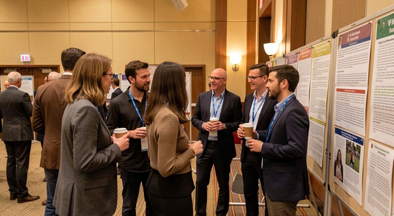 Researchers of several nationalities talking in small groups beside poster boards in a conference hall between sessions, lanyards and coffee cups in hand, warm interior light.